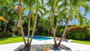 Ein Poolbereich mit freiem Blick auf den Pool, umgeben von Palmen und Grünpflanzen. Im Vordergrund stehen Korbstühle und ein Blumentopf.