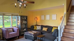 A living room with a sofa, armchair, and ottoman, a bookshelf, a ceiling fan, and a staircase. - Charming 3-bedroom riverside cottage in brilliant Tawas City (Tawas City)