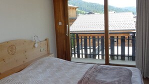 A bedroom with a wooden headboard, a bed with a patterned bedspread, and a view of a balcony with a wooden railing and a mountain backdrop. - Spacious 2-Room Apt for 4 in Les Gets Center - Wifi, Linens Included, Near Shops & Ski Lifts (Les Gets)