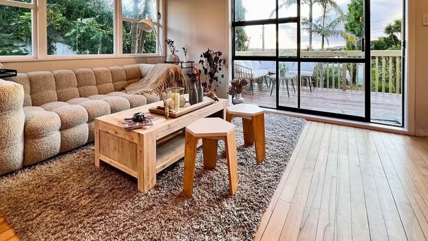 A spacious living room with a large window, a wooden coffee table, a beige sofa, and a view of a deck and palm trees. - Guest Favorite 3BRM - New Reno with Parking & AC - CBD 12KMs & AP 17KMs  (Auckland)
