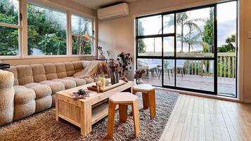 A spacious living room with a large window, a wooden coffee table, a beige sofa, and a view of a deck and palm trees.