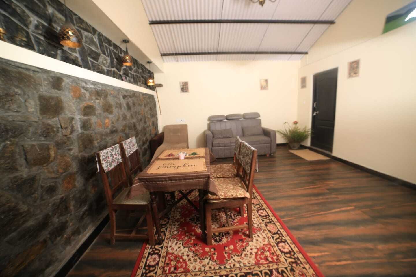 A dining area with a long wooden table, six chairs, a red patterned rug, a stone wall, and a grey sofa in the background.