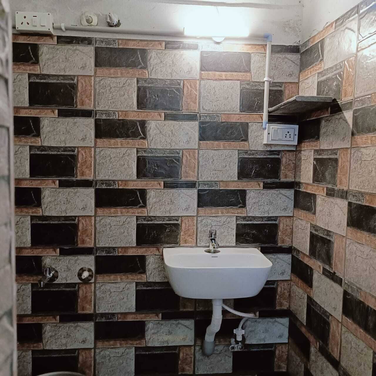 Un baño con lavamanos blanco, paredes cubiertas de azulejos en varios tonos de marrón y negro, y tuberías a la vista.