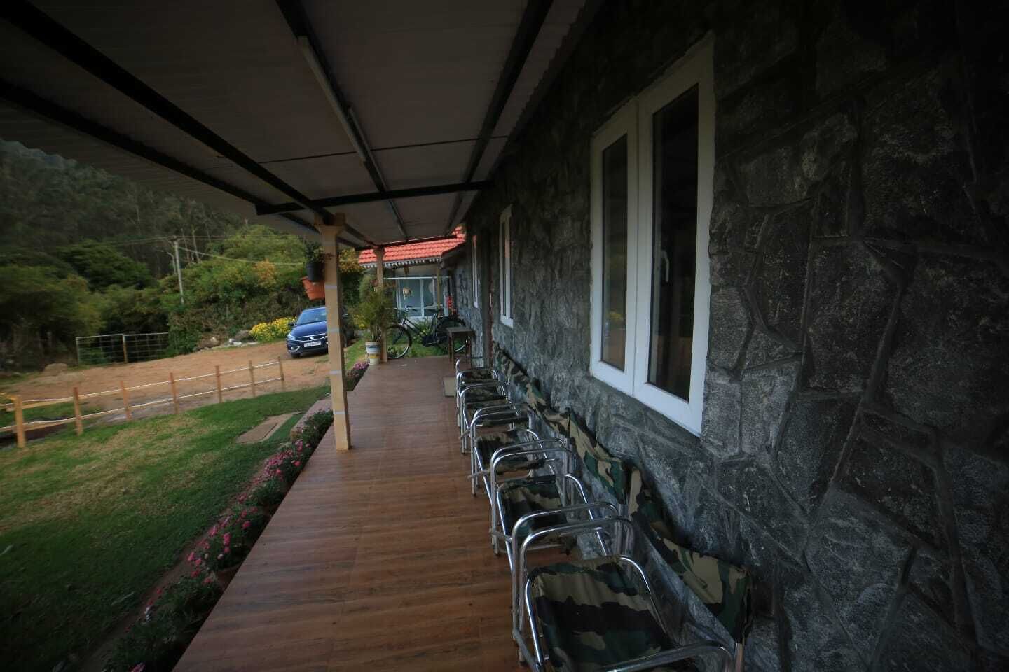 A covered outdoor seating area with metal chairs, a stone building with windows, and a pathway leading to a parked car.