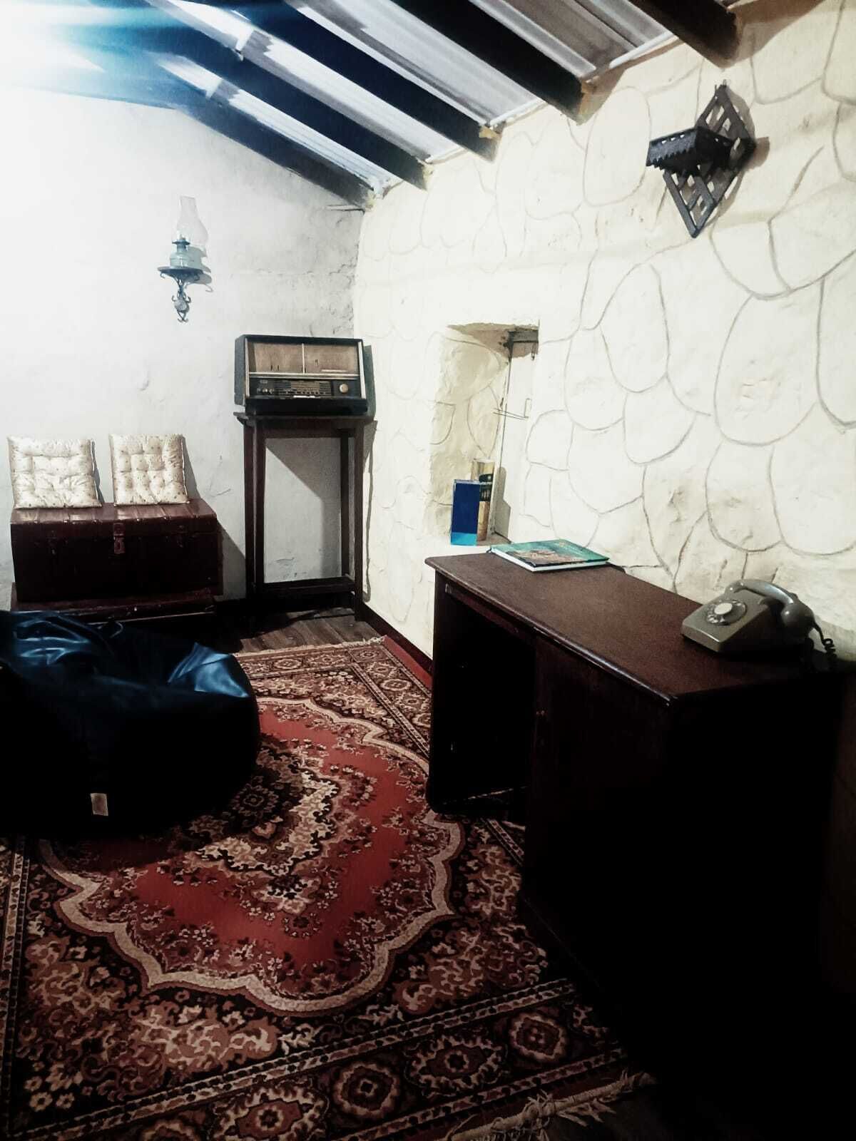 A room with a patterned rug, a wooden desk with a telephone, a radio, and a wall-mounted light fixture.