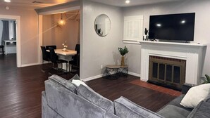 Un salon moderne avec un canapé gris, un téléviseur à écran plat, une cheminée et un coin repas comprenant une table ronde et des chaises.