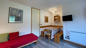 Una habitación compacta con un sofá rojo, una pequeña mesa de comedor con sillas, un televisor montado en la pared y una puerta que da a otra habitación.