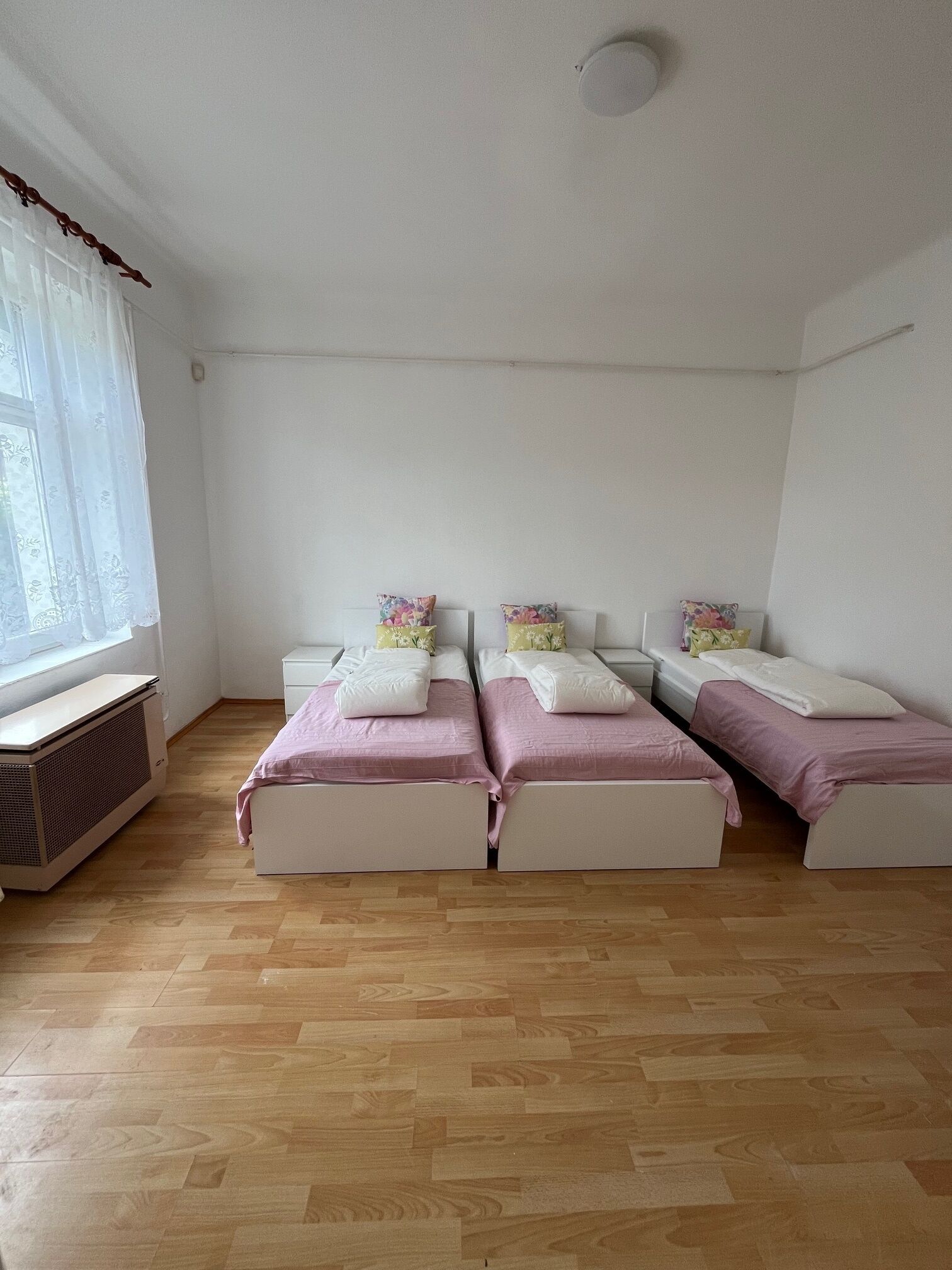 Un dormitorio con dos camas, una ventana con cortinas translúcidas, un radiador y un techo blanco.