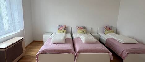 Un dormitorio con dos camas, una ventana con cortinas translúcidas, un radiador y un techo blanco.