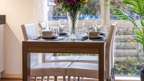 Mesa de comedor para cuatro personas, con un jarrón de flores, sillas blancas y vistas a árboles a través de una ventana.