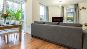 Una sala de estar moderna con un sofá gris, piso de madera, una mesa de comedor con sillas, un televisor mostrando 'NETFLIX' y ventanas amplias con cortinas.
