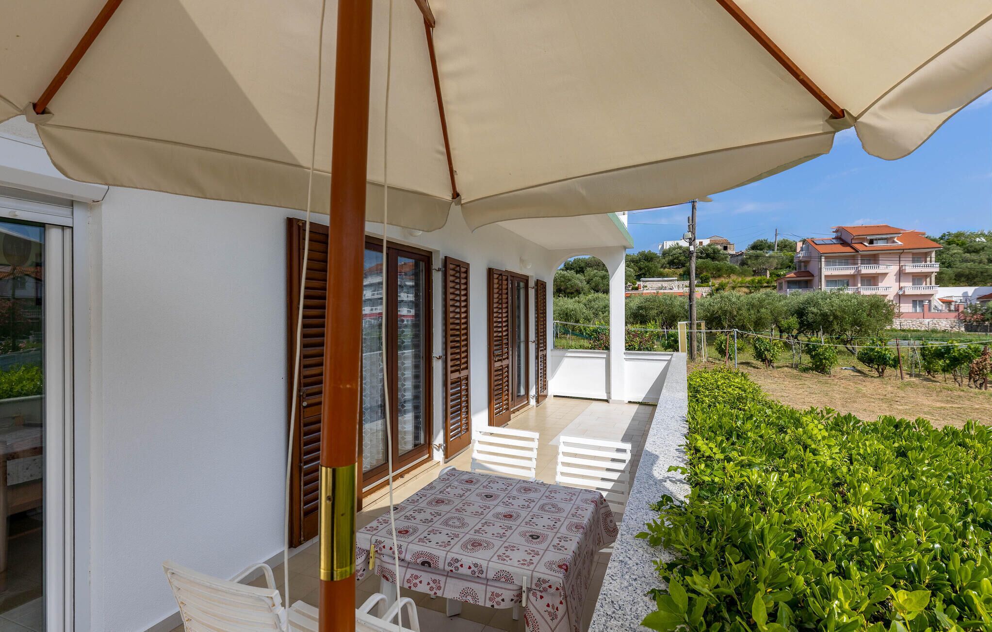 A balcony with a table and chairs, wooden shutters, and a view of a residential area with houses and greenery.