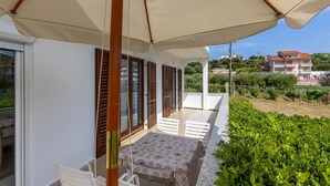 A balcony with a table and chairs, wooden shutters, and a view of a residential area with houses and greenery.