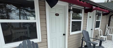 A covered porch with Adirondack chairs, a white door with a number plate, and a window with a view of trees.