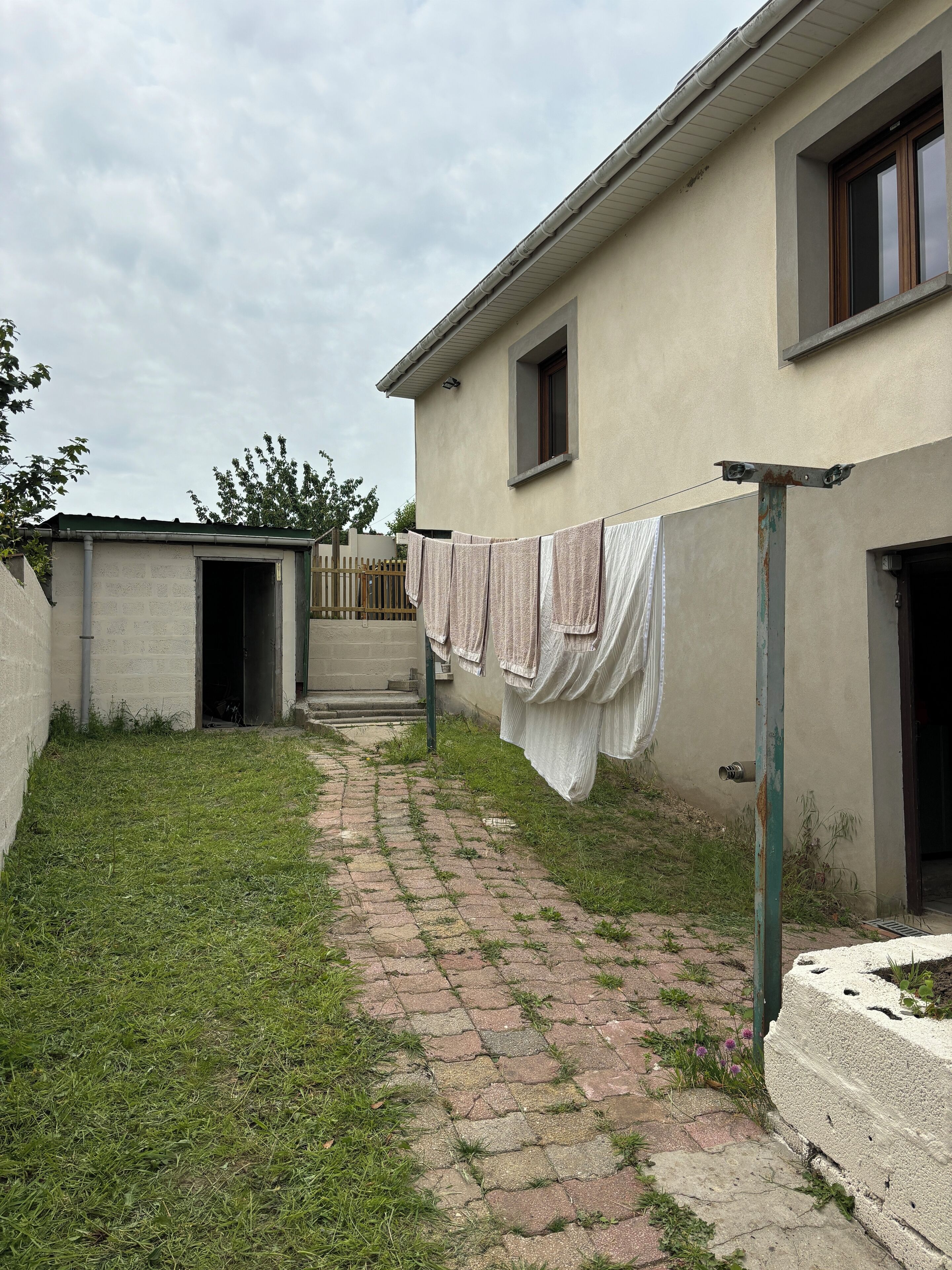 Un edificio con un camino pavimentado que lleva a una puerta, un tendedero con ropa y una zona de césped.