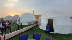 Une rangée de tentes en forme de dôme, séparées par une zone herbeuse, avec une rambarde et quelques chaises.