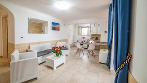 Een woonkamer met een bank, salontafel en een eethoek met stoelen en een kleine keuken.