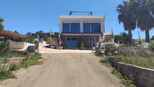 Une maison à deux étages avec une entrée centrale, des piliers en pierre et un jardin agrémenté de palmiers et de fleurs.