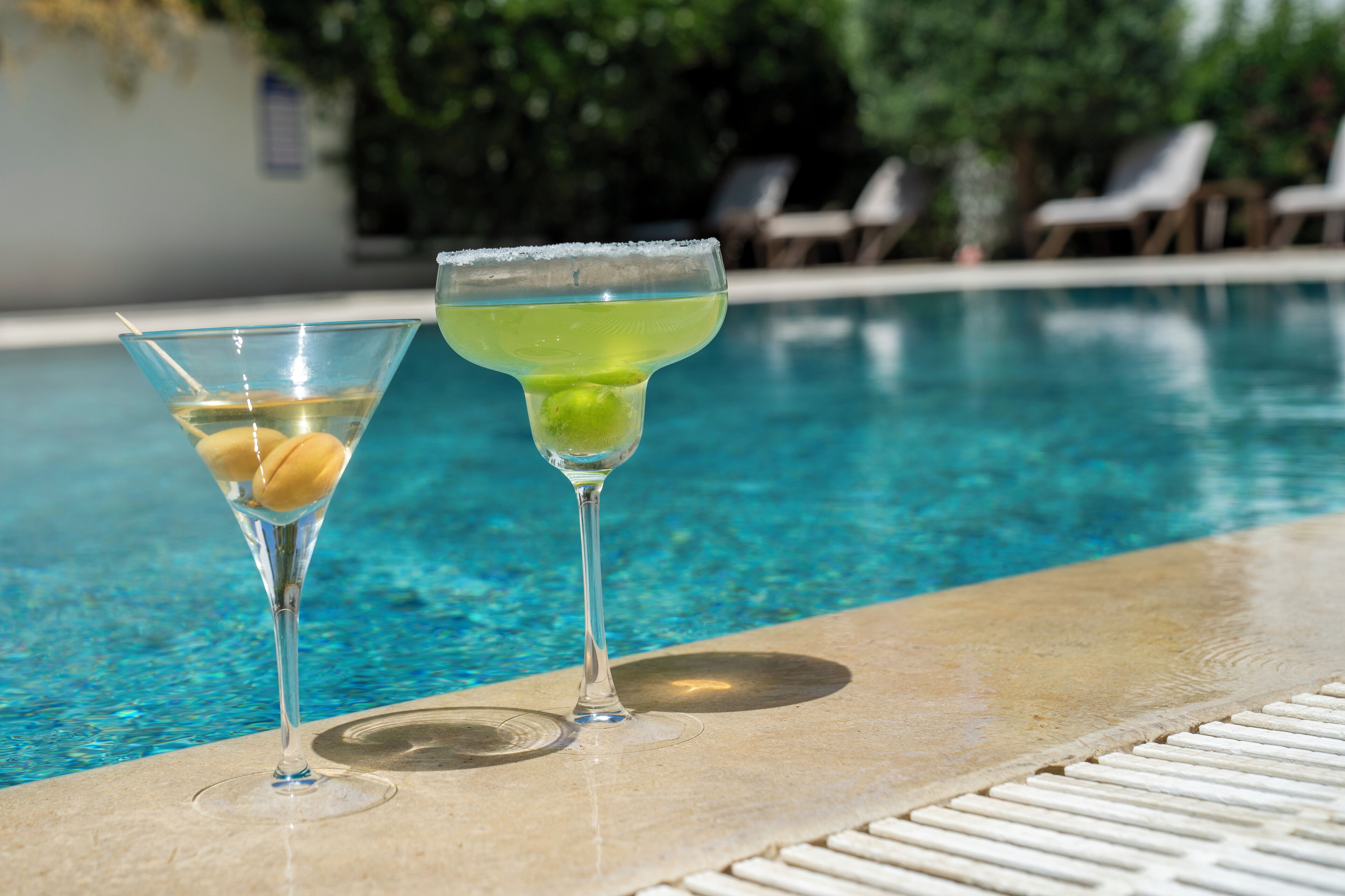 Two martini glasses on a poolside with clear blue water.