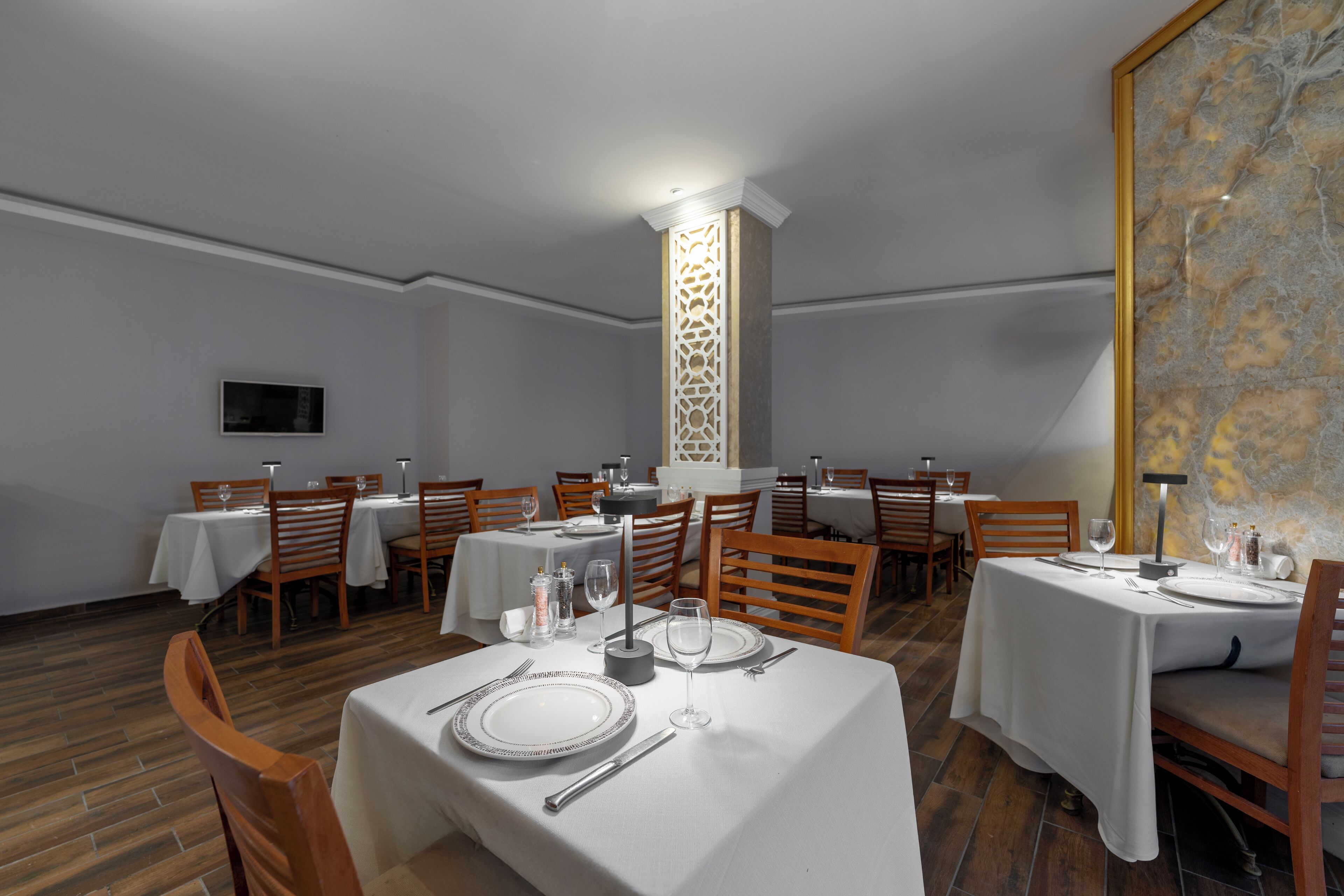 Uma sala de jantar com mesas postas para quatro pessoas, cadeiras de madeira e um pilar decorativo.