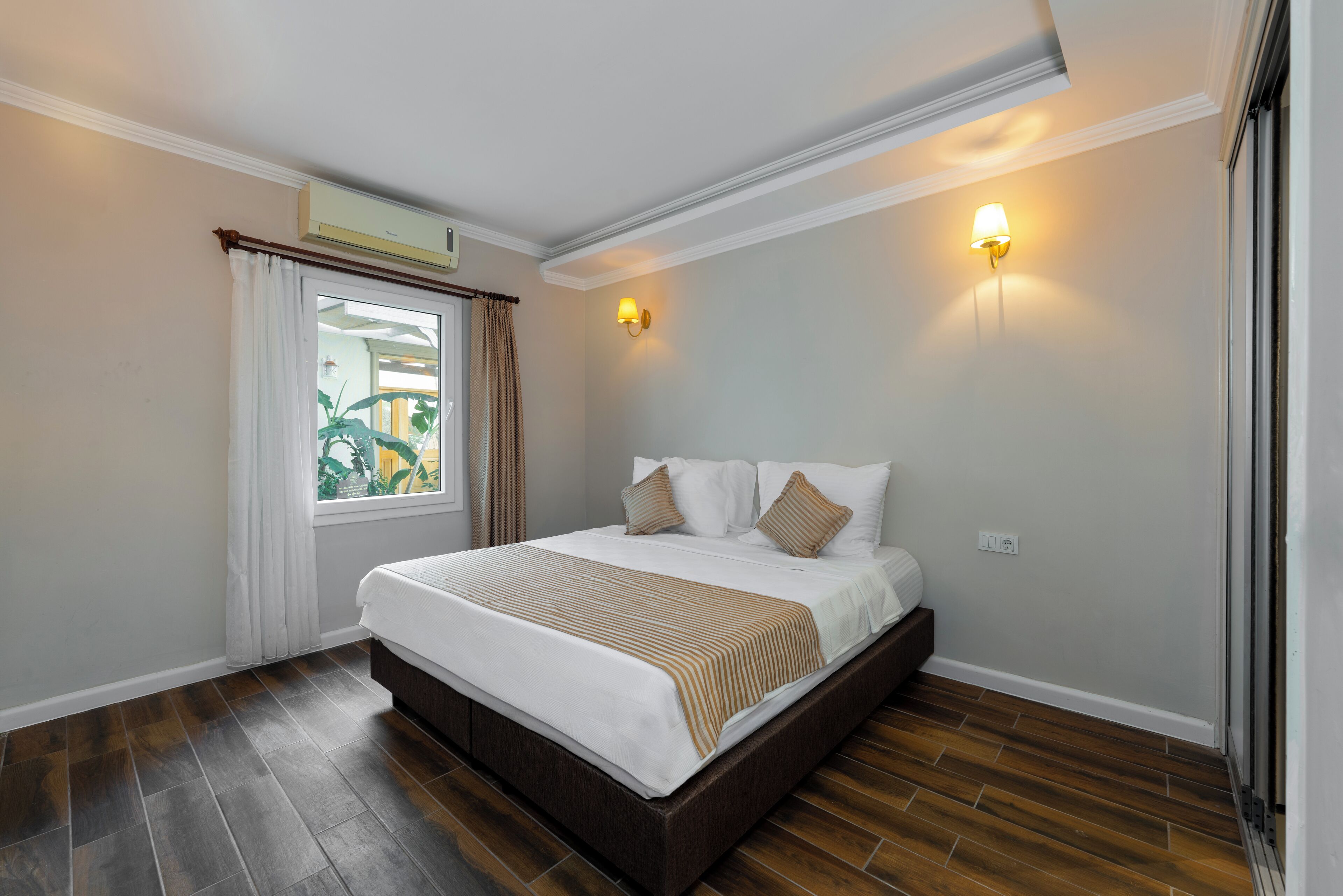 Un lit bien fait, recouvert d’une couverture rayée blanche et beige, une fenêtre avec des rideaux et un luminaire fixé au mur.