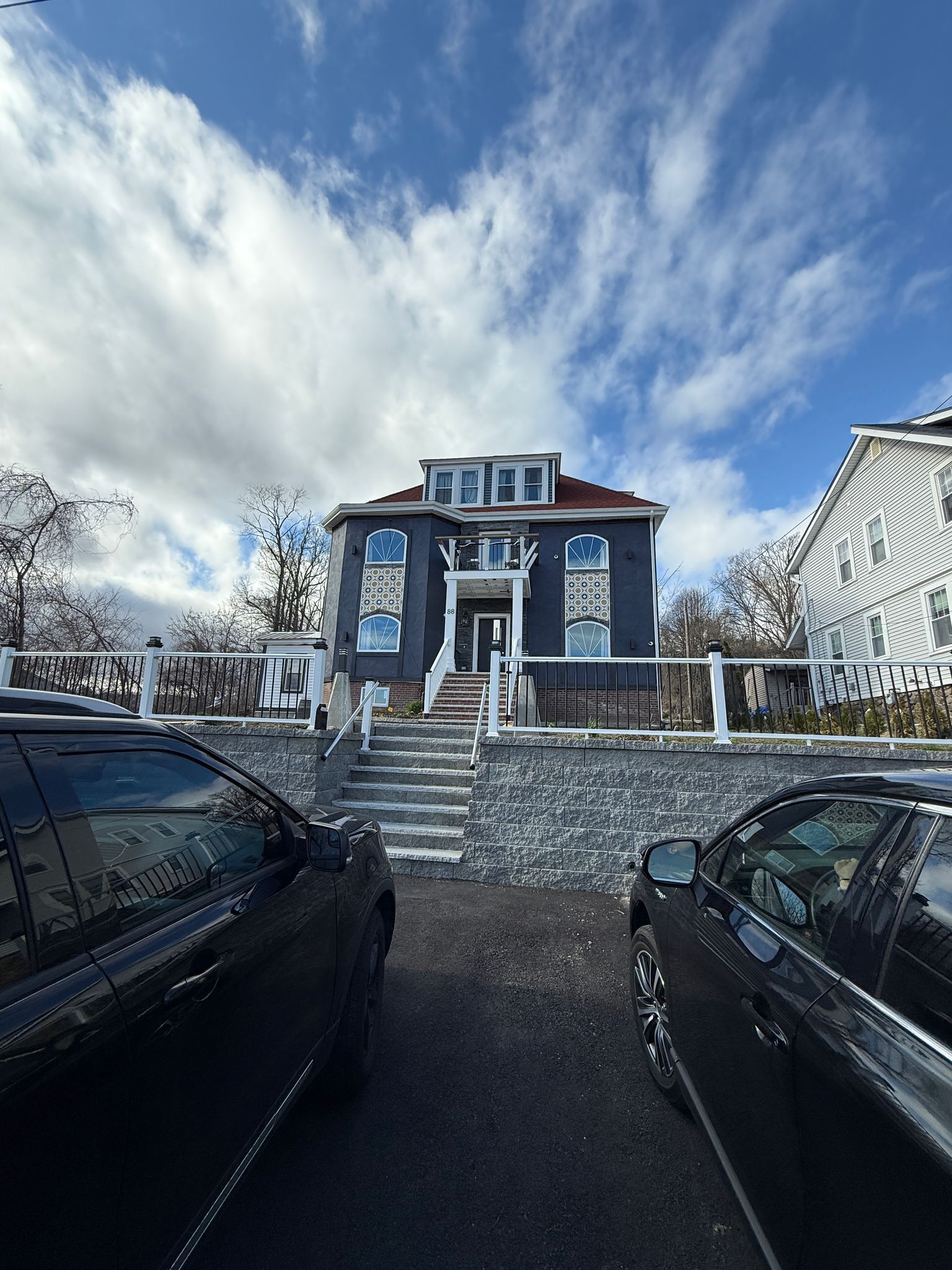 A two-story building with a central entrance, a staircase leading to the entrance, and a parking area with two cars.