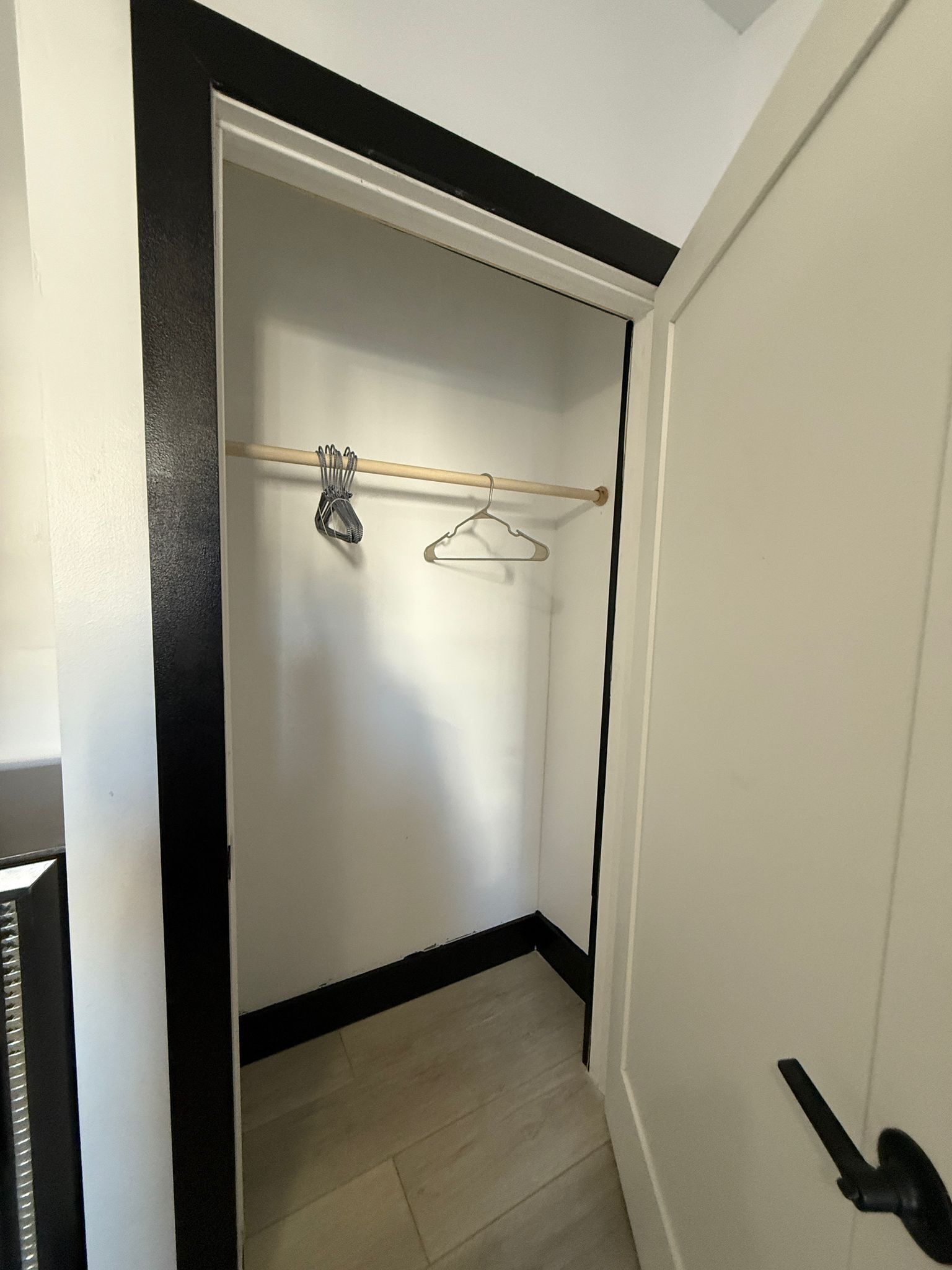 A narrow hallway with a wooden floor, a white door on the right, and a coat rack with hangers and a broom in the center.