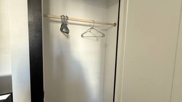 A narrow hallway with a wooden floor, a white door on the right, and a coat rack with hangers and a broom in the center.