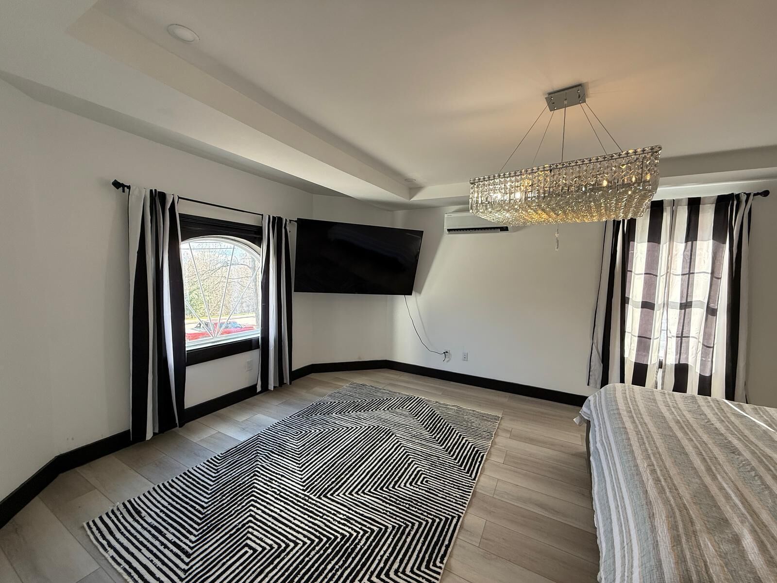 A modern bedroom with a large bed, a black and white patterned rug, a chandelier, and a TV mounted on the wall.