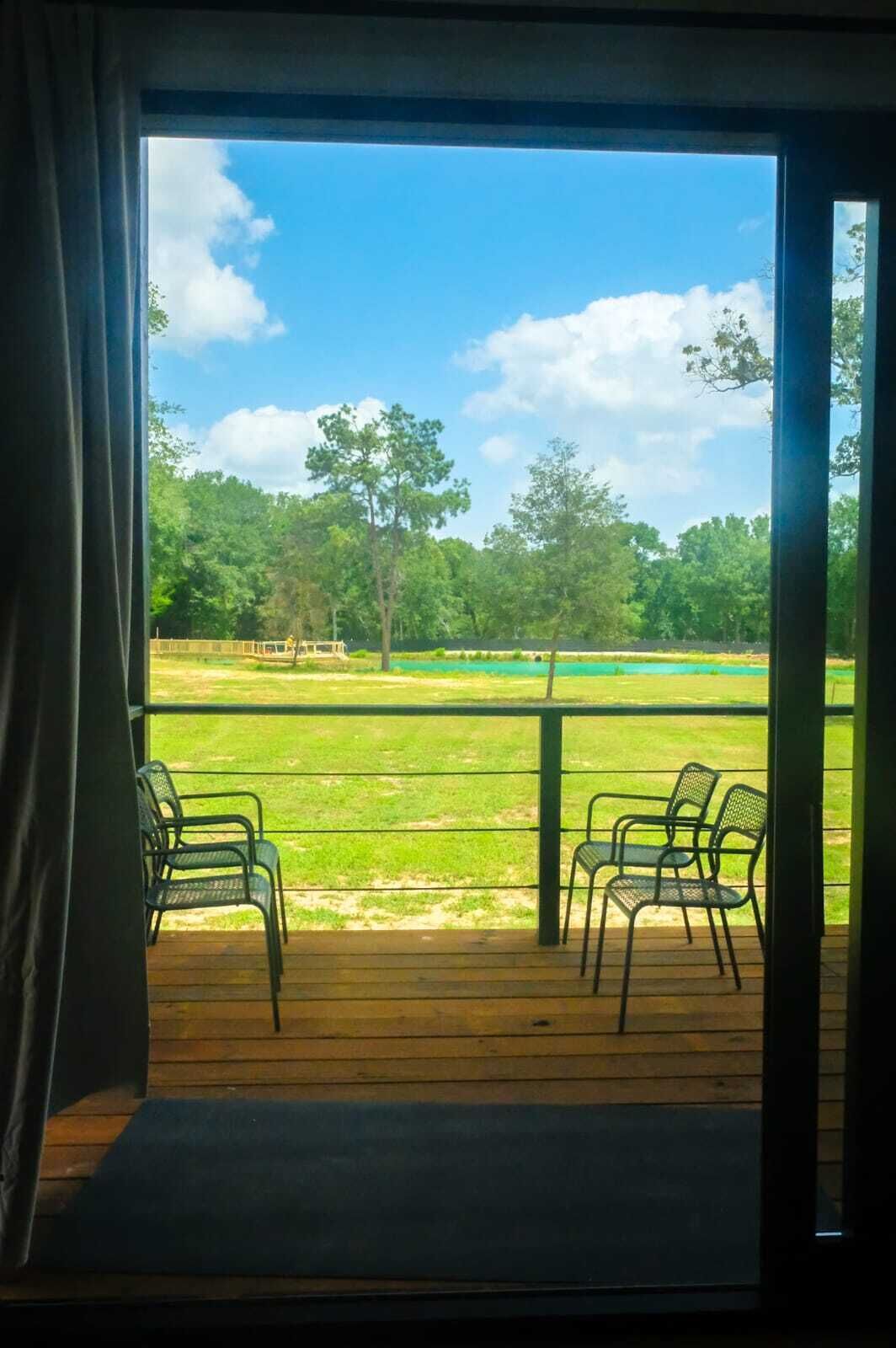 Una habitación con un gran ventanal corredero que da a un espacio exterior con césped y árboles. Hay dos sillas en el porche.