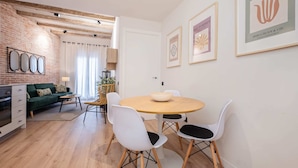 A modern living room with a green sofa, a coffee table, and a dining area with a round table and white chairs. There are framed artworks on the wall and a brick wall with exposed beams.