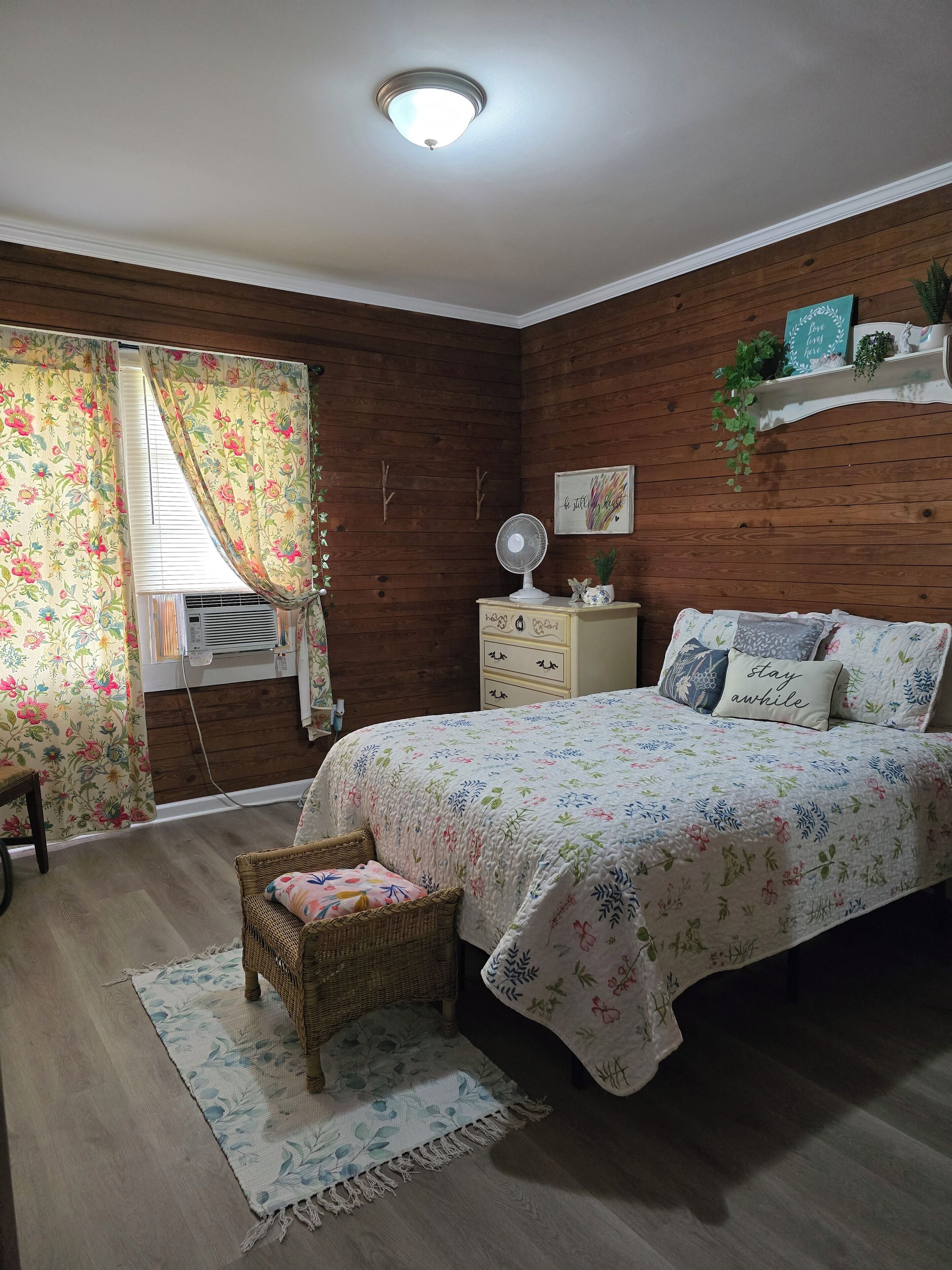 A bedroom with a wooden wall, a bed with floral bedding, a bedside table, a wall-mounted fan, and a window with floral curtains.