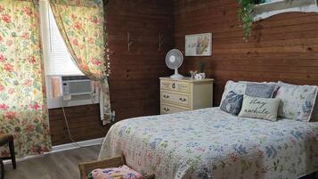 A bedroom with a wooden wall, a bed with floral bedding, a bedside table, a wall-mounted fan, and a window with floral curtains.