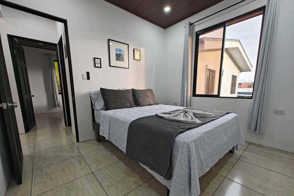 A neatly arranged bedroom with a bed, a window with curtains, and a door leading to another room.