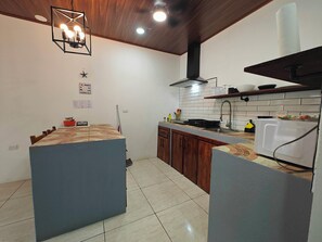 A kitchen with a central island, wooden cabinets, a tiled backsplash, and a modern pendant light. - AJ House & Suites (Bahía Ballena)