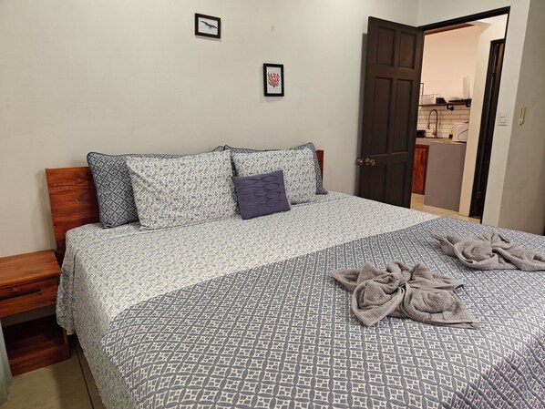 A neatly made bed with patterned bedding, a wooden headboard, and a door leading to a kitchen area. - AJ House & Suites (Bahía Ballena)
