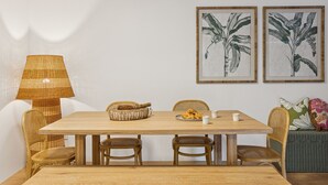 A dining area with a wooden table, wicker chairs, a woven lamp, and two framed botanical prints on the wall.