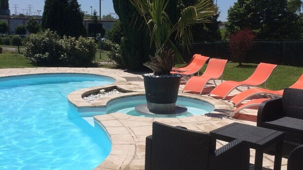 Une piscine bordée d’une terrasse en pierre, entourée de chaises longues orange et d’une table en osier noir.