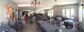 A spacious dining area with multiple tables and chairs, a kitchen in the background, and a large painting on the wall. - Balladins Lyon Est (Saint-Quentin-Fallavier)