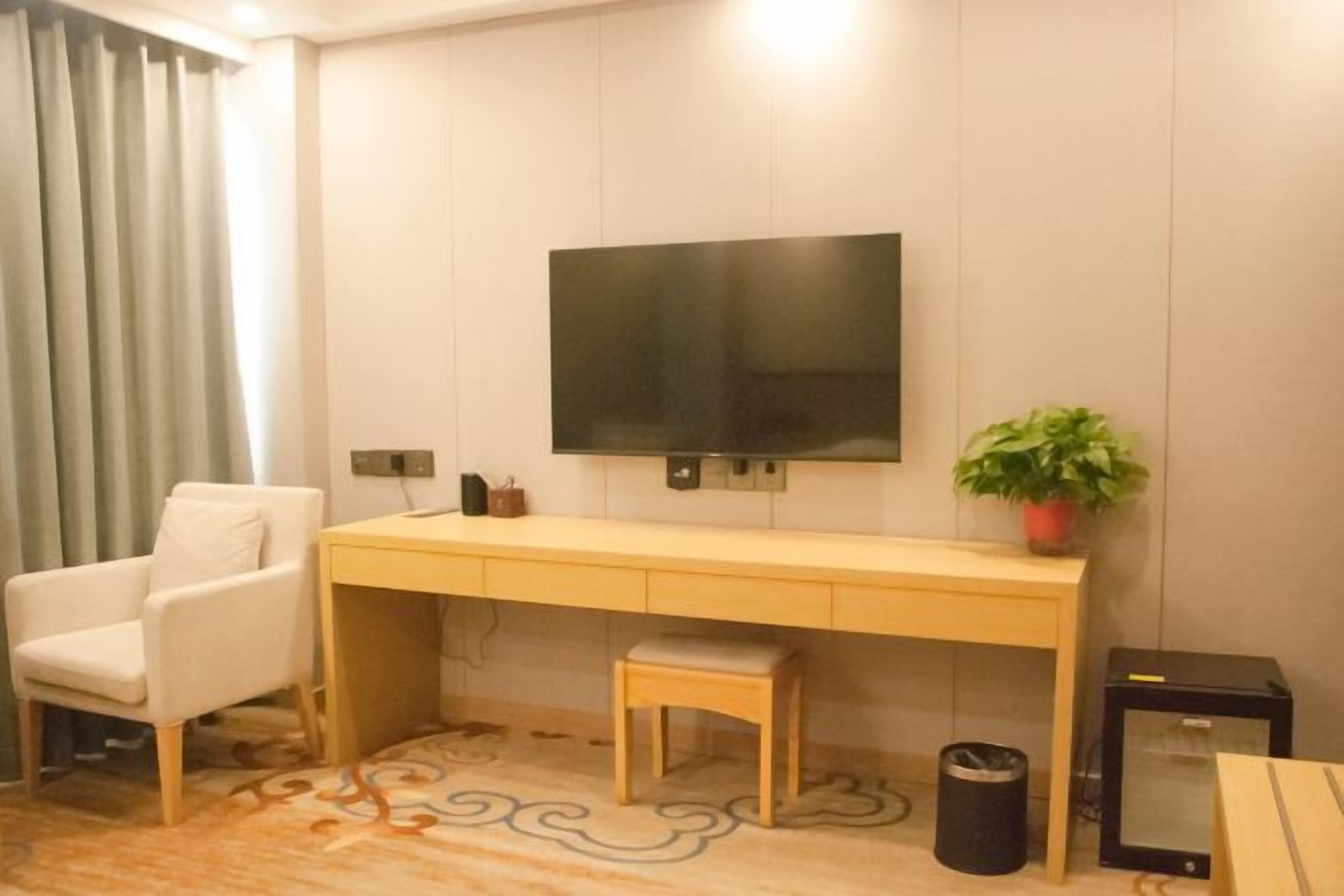 Une chambre moderne avec un bureau en bois, un téléviseur fixé au mur, une chaise blanche, un petit tabouret, une plante en pot et une poubelle.