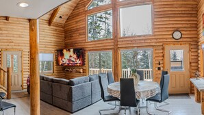 Ein gemütliches Blockhaus-Interieur mit Kamin, einer Couch, einem Esstisch mit Stühlen und großen Fenstern.