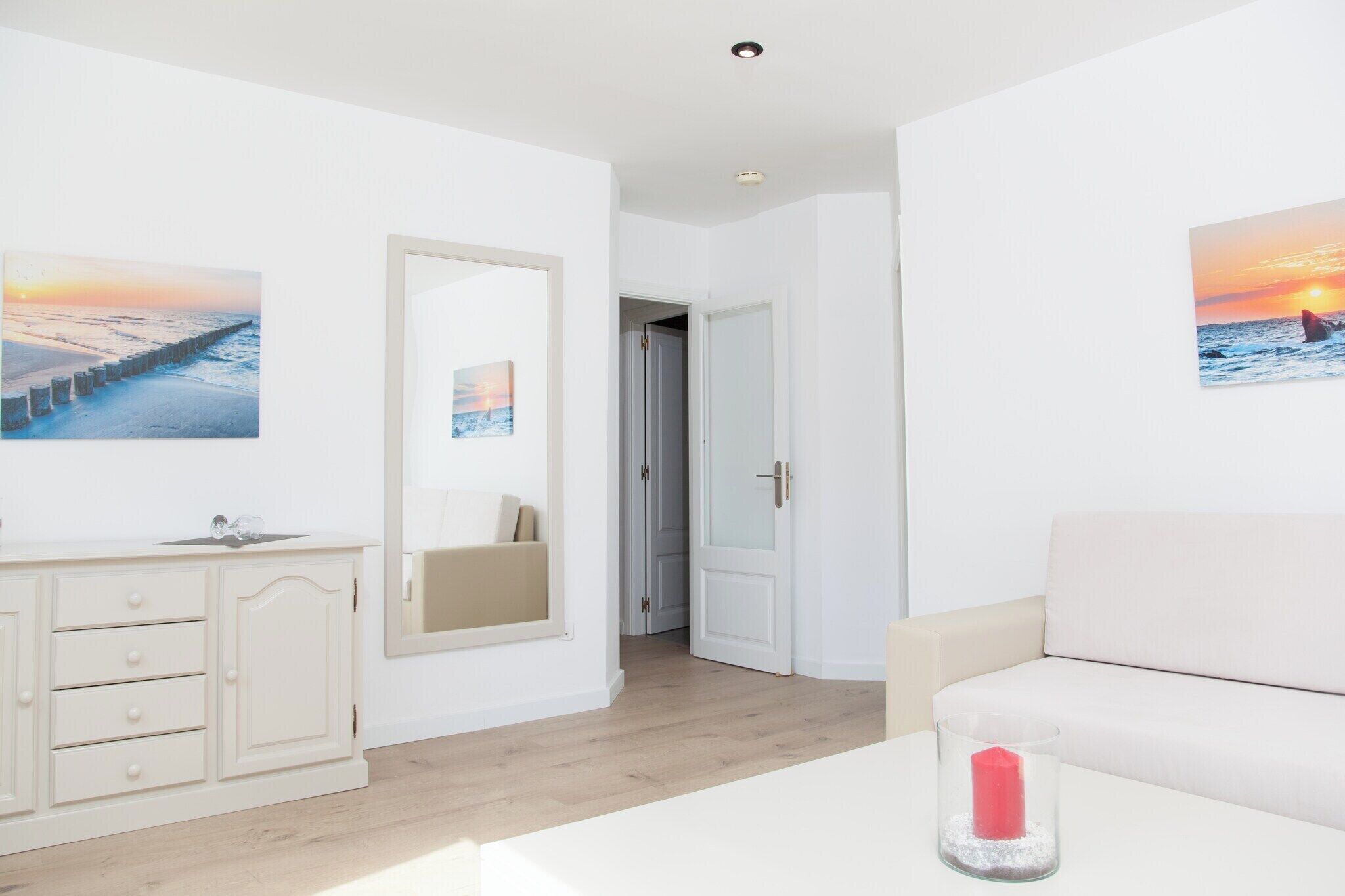 Une pièce moderne et minimaliste, avec un canapé blanc, une table en verre sur laquelle est posée une bougie, un meuble blanc et deux tableaux encadrés représentant un coucher de soleil sur l’océan.
