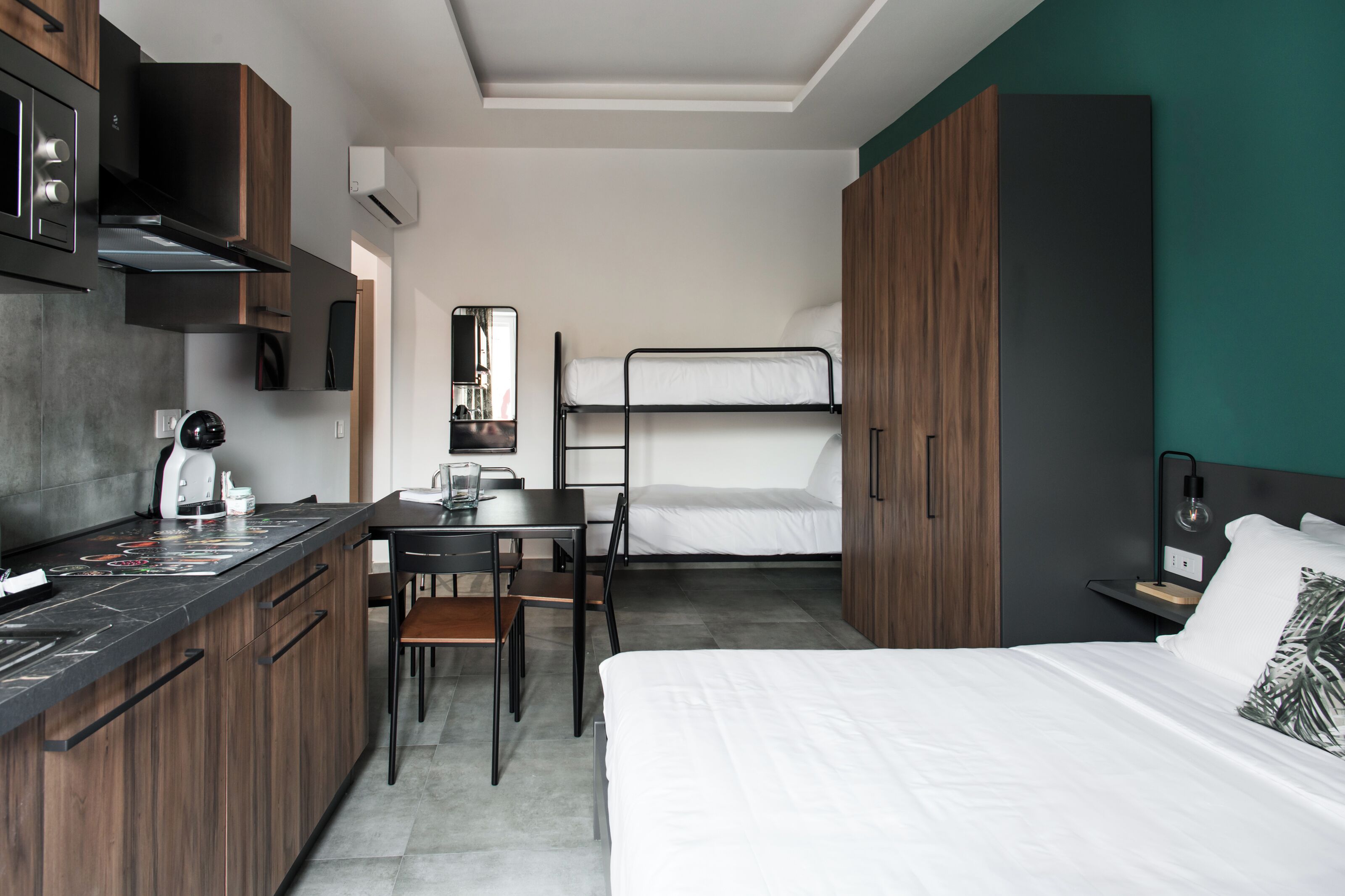 A compact hotel room with a bunk bed, a kitchenette with a coffee machine, a dining table, and a mirror.