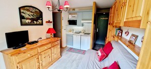 Ein kompakter Wohnbereich mit Fernseher, Holzschränken, einer roten Lampe, einer Couch mit roten Kissen und einer Küchenzeile mit Mikrowelle und Schränken.