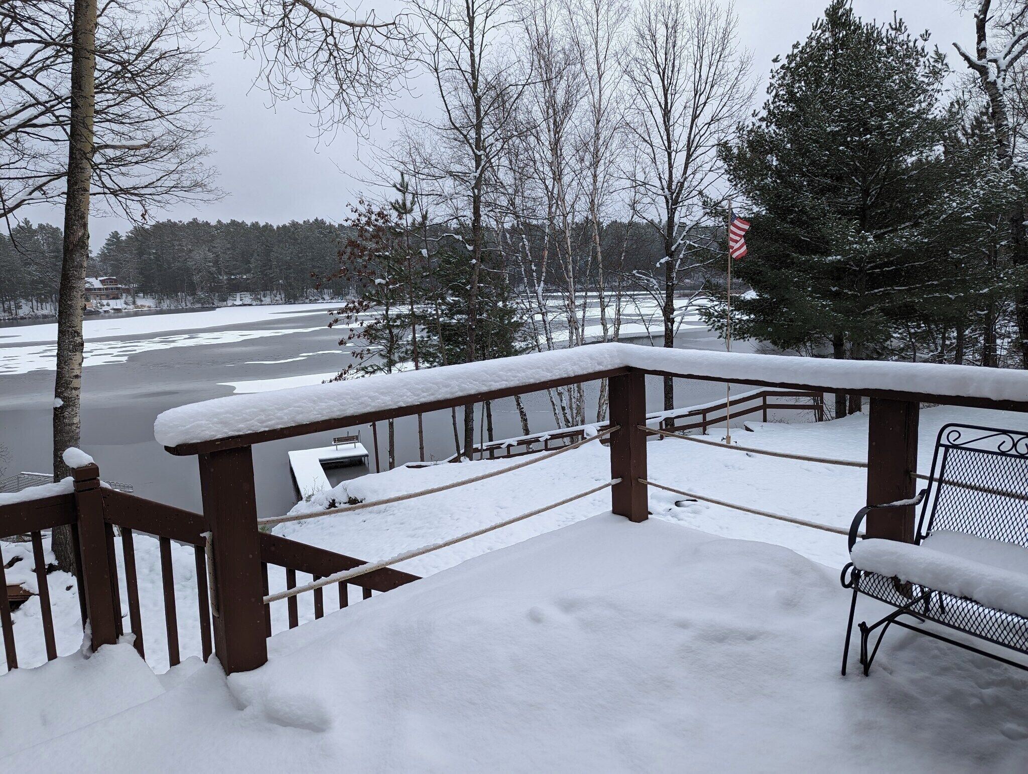 A snow-covered deck with a bench and a railing overlooking a frozen lake and trees.