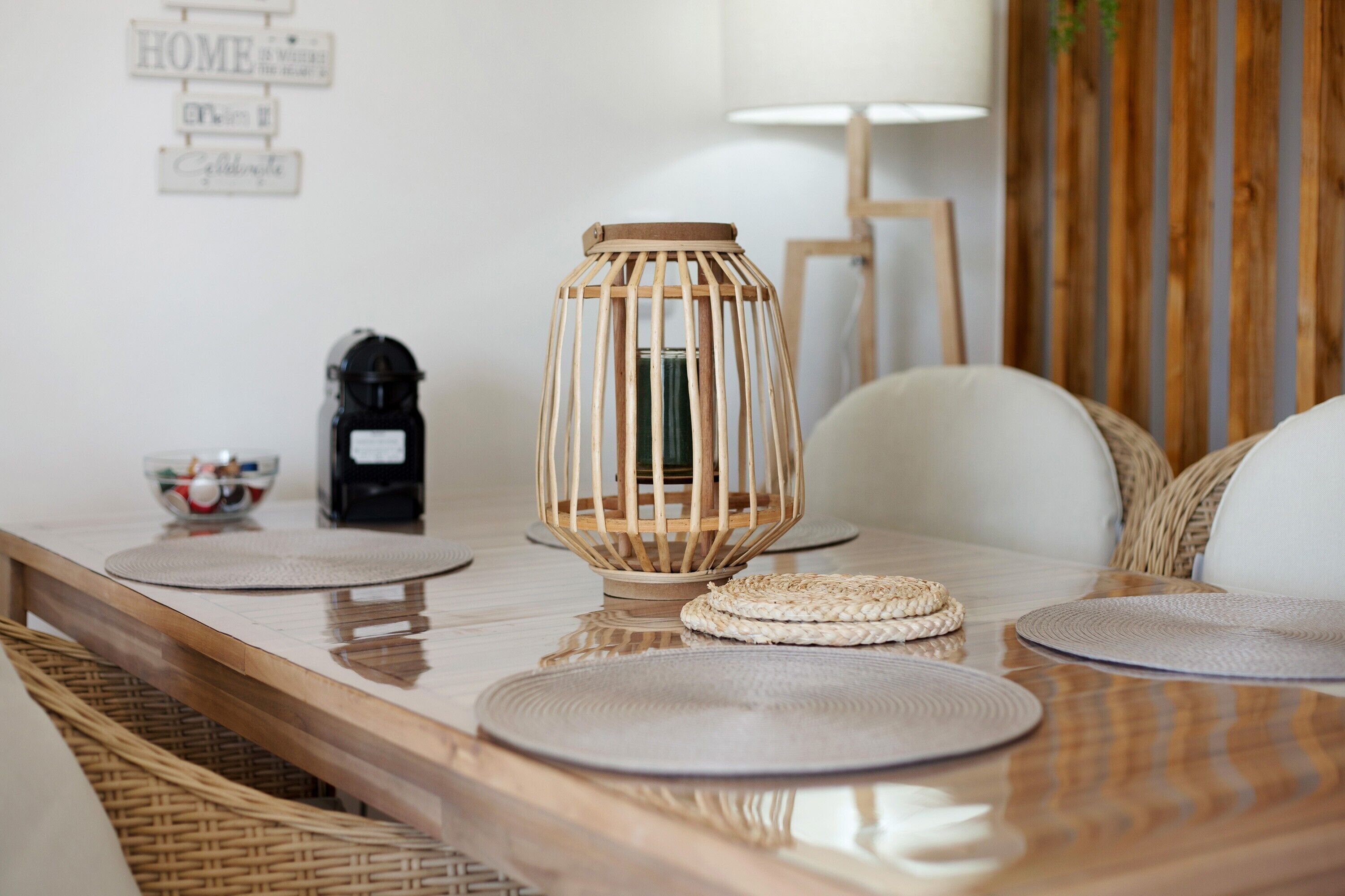 A wooden dining table with woven placemats, a woven basket, a wooden lantern, and a coffee machine.