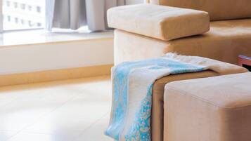Un canapé beige surmonté d’un plaid à motifs bleus, une fenêtre avec des rideaux et un sol carrelé.
