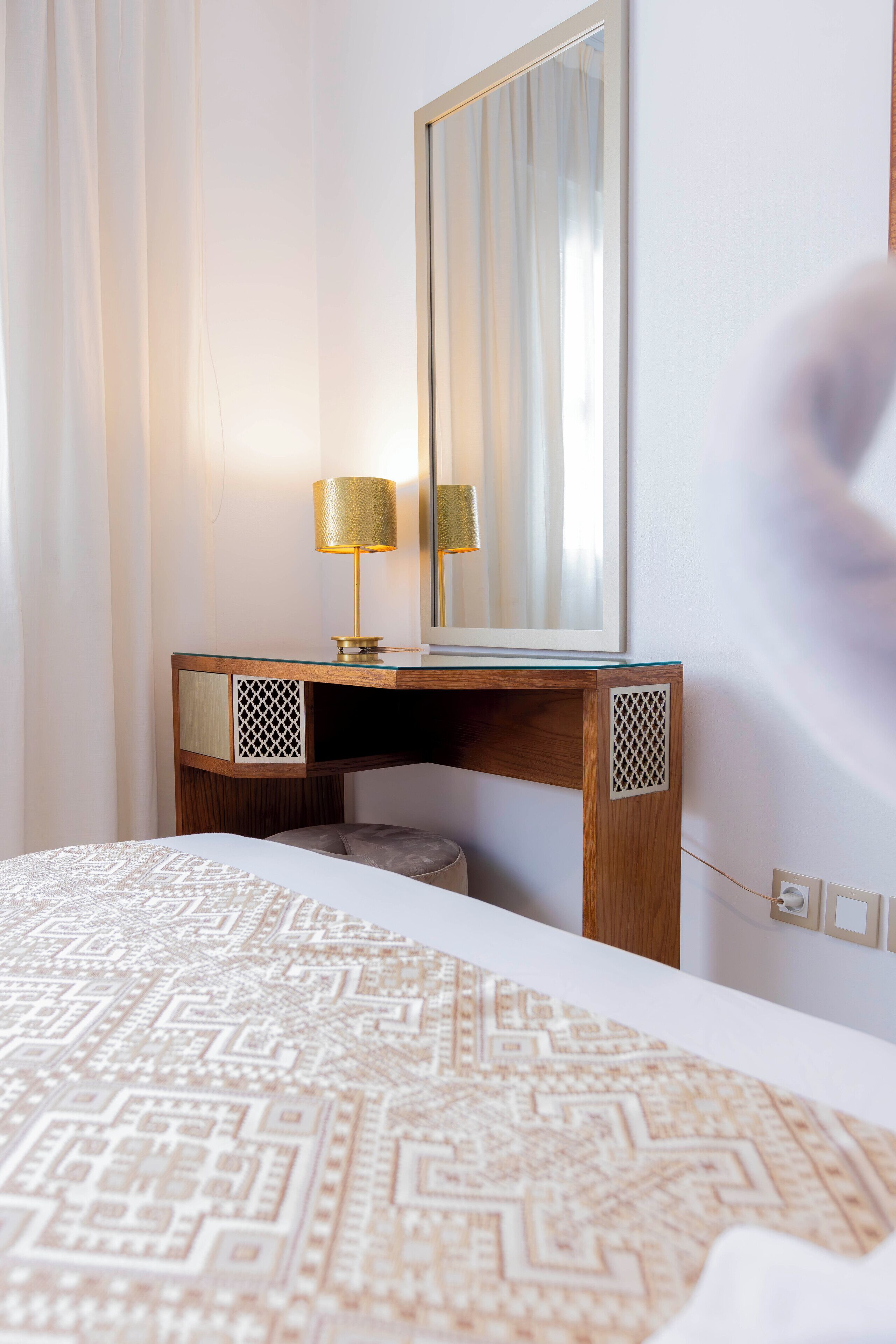 A hotel room with a bed, a wooden desk with a lamp, a mirror, and a patterned bedspread.