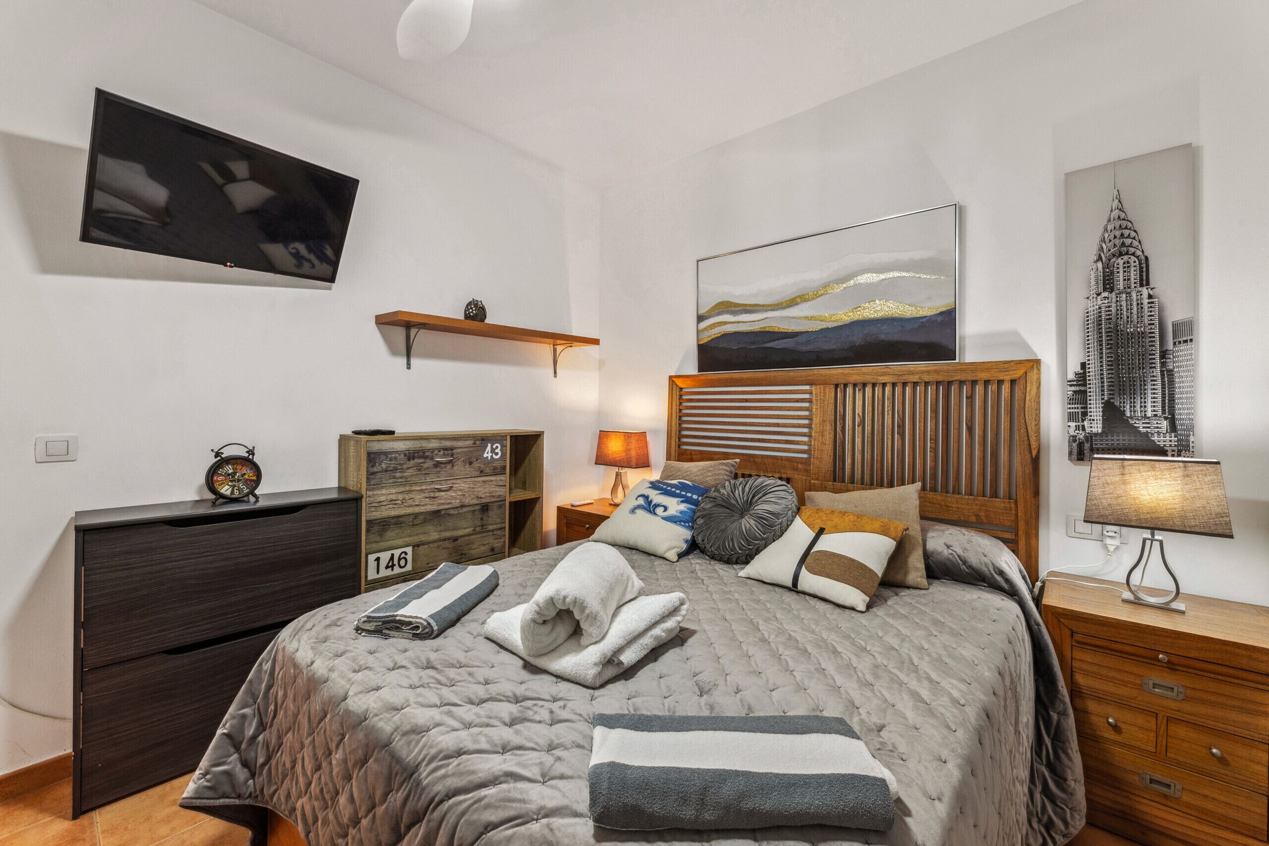 Une chambre à coucher bien rangée, avec un lit, une commode, un téléviseur fixé au mur et un tableau représentant un paysage urbain.