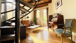 Un salon spacieux avec un escalier en bois, un piano et une grande fenêtre qui laisse entrer la lumière naturelle.
